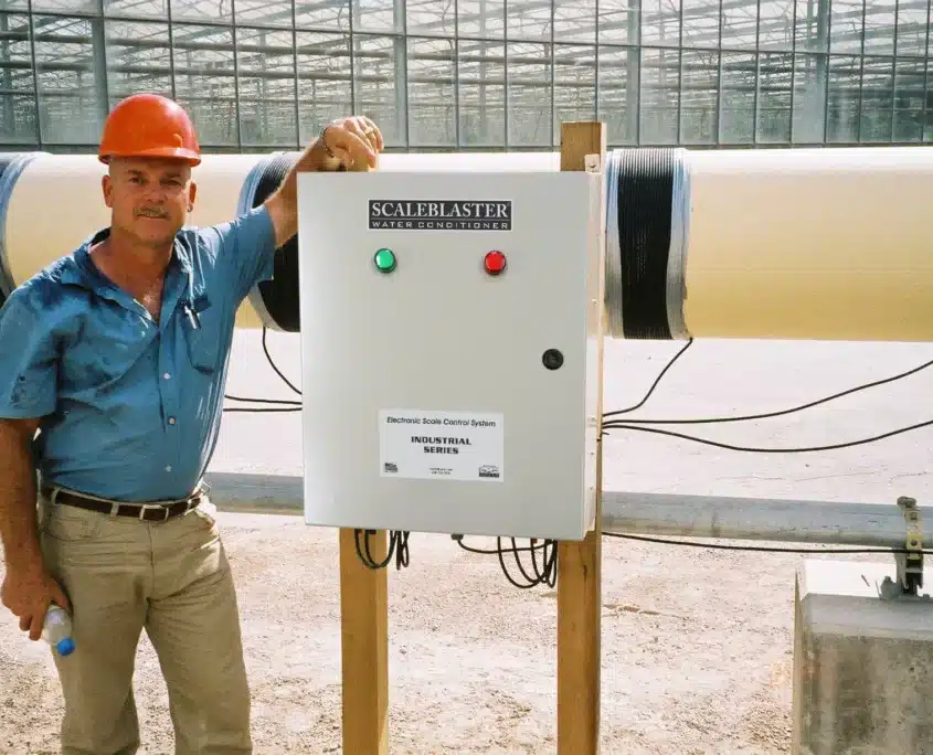Technician standing beside a ScaleBlaster industrial water conditioner installed on large piping.