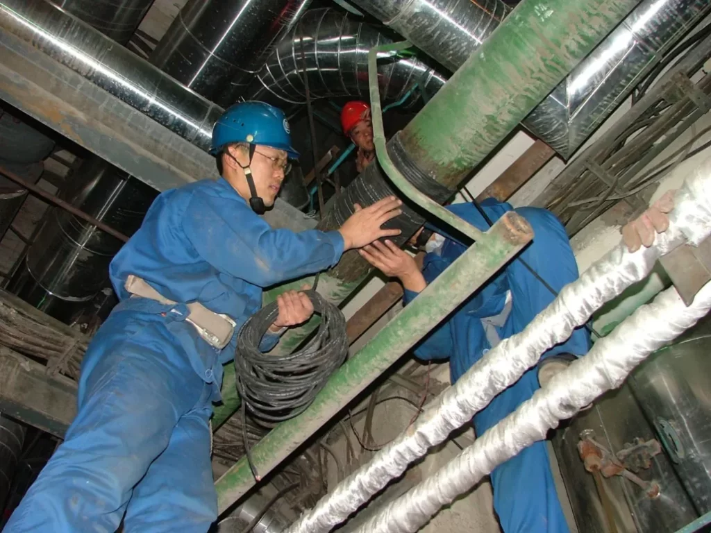 Workers installing ScaleBlaster cables on industrial pipes in a mechanical room or plant setting.