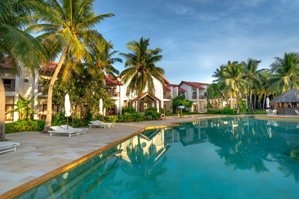 Resort swimming pool surrounded by palm trees, lounge chairs, and white buildings.